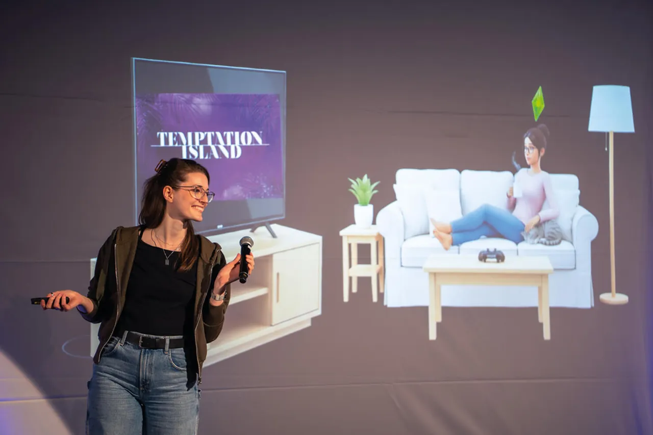 Eine junge Frau mit Brille hält ein Mikrofon in der Hand und gestikuliert während eines Vortrags. Im Hintergrund wird auf einer Leinwand ein Wohnzimmer mit zwei Personen gezeigt, die auf einem Sofa sitzen.