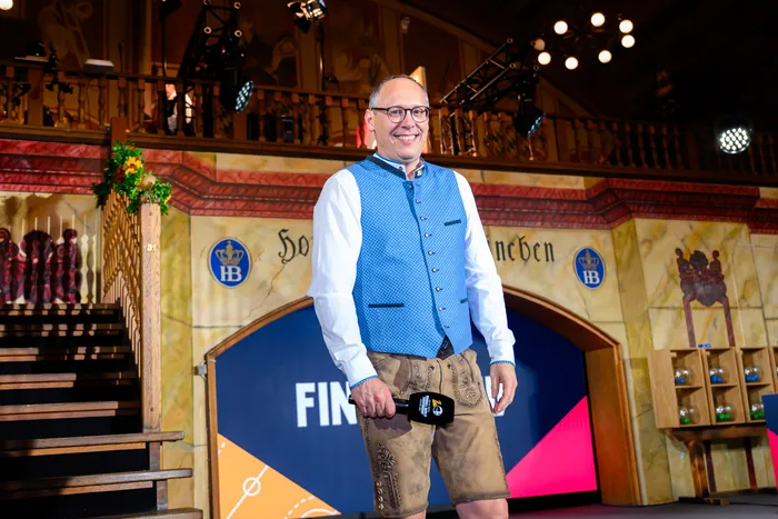 Ein Mann mit Brille, im karierten Hemd und Lederhose steht lächelnd auf einer Bühne vor einem Hintergrund, der an ein bayerisches Brauhaus erinnert. Er hält ein Mikrofon in der Hand.