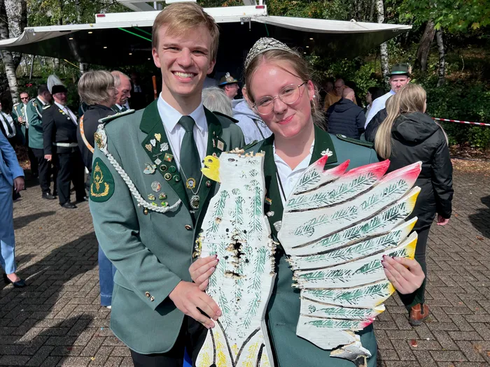 Ein junger Mann und eine junge Frau in grünen Uniformen mit Orden halten eine große, bemalte Holzfigur eines Vogels.