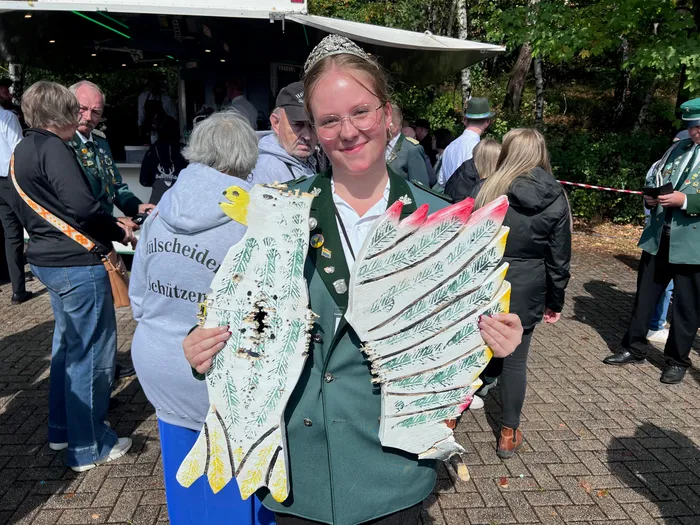 Eine junge Frau mit Tiara und grüner Uniform hält einen großen, weiß-grün bemalten Holzvogel.