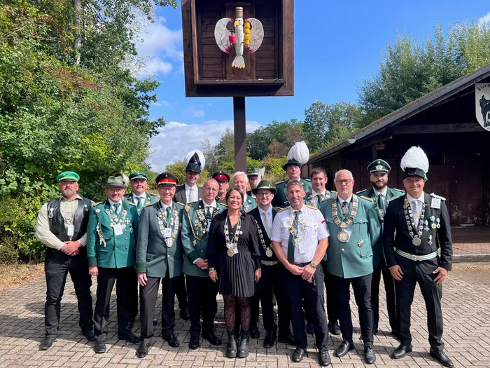 Eine Gruppe von Personen in traditionellen Uniformen mit Orden und Federhüten posiert beim Kreiskönigsschießen vor einem Holzgebäude.