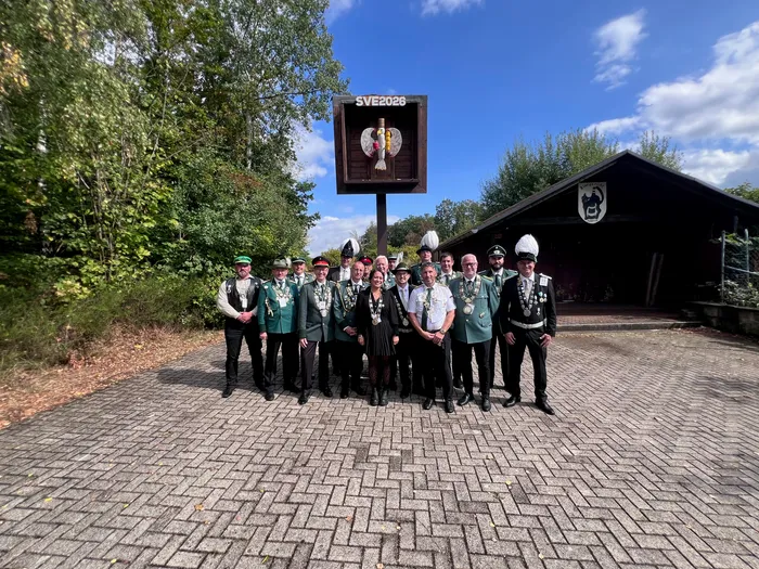 Eine Gruppe von Schützen in grünen Uniformen mit Orden und weißen Hüten steht vor einem Holzgebäude und einem Schild mit der Aufschrift „SVE 2026“.