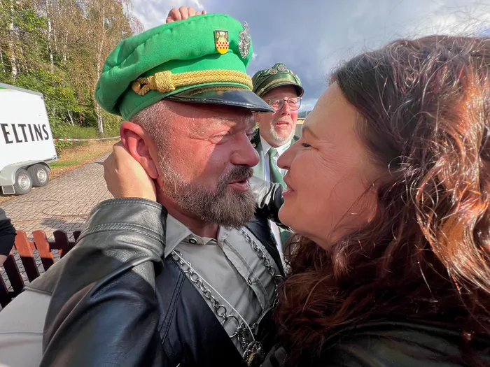 Ein Mann mit grünem Schützenhut wird von einer Frau umarmt, im Hintergrund steht ein weiterer Mann in Schützenuniform.