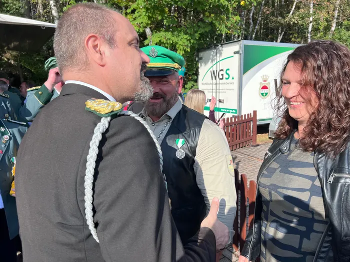 Ein Mann in dunkler Uniform mit silbernen Schulterstücken unterhält sich mit einem bärtigen Mann in grüner Kopfbedeckung und einer lächelnden Frau.