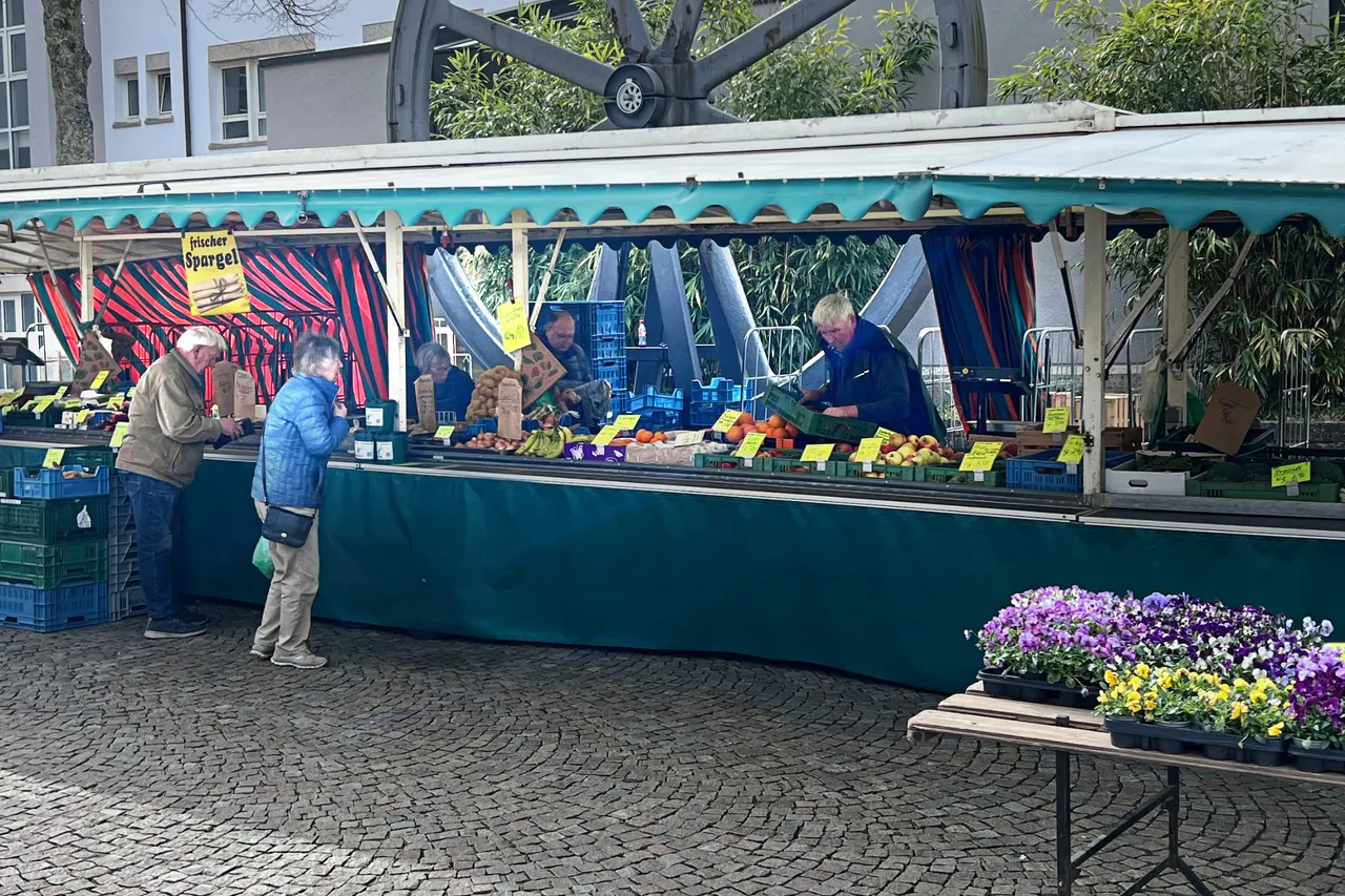 Gemüse- und Obsthändler Günter Deitelhoff bedient Kunden an seinem Marktstand in Plettenberg. Im Hintergrund steht seine Frau Heiderose. Vor dem Stand stehen Kisten mit Kartoffeln, Obst und Gemüse und ein Tisch mit blühenden Pflanzen.