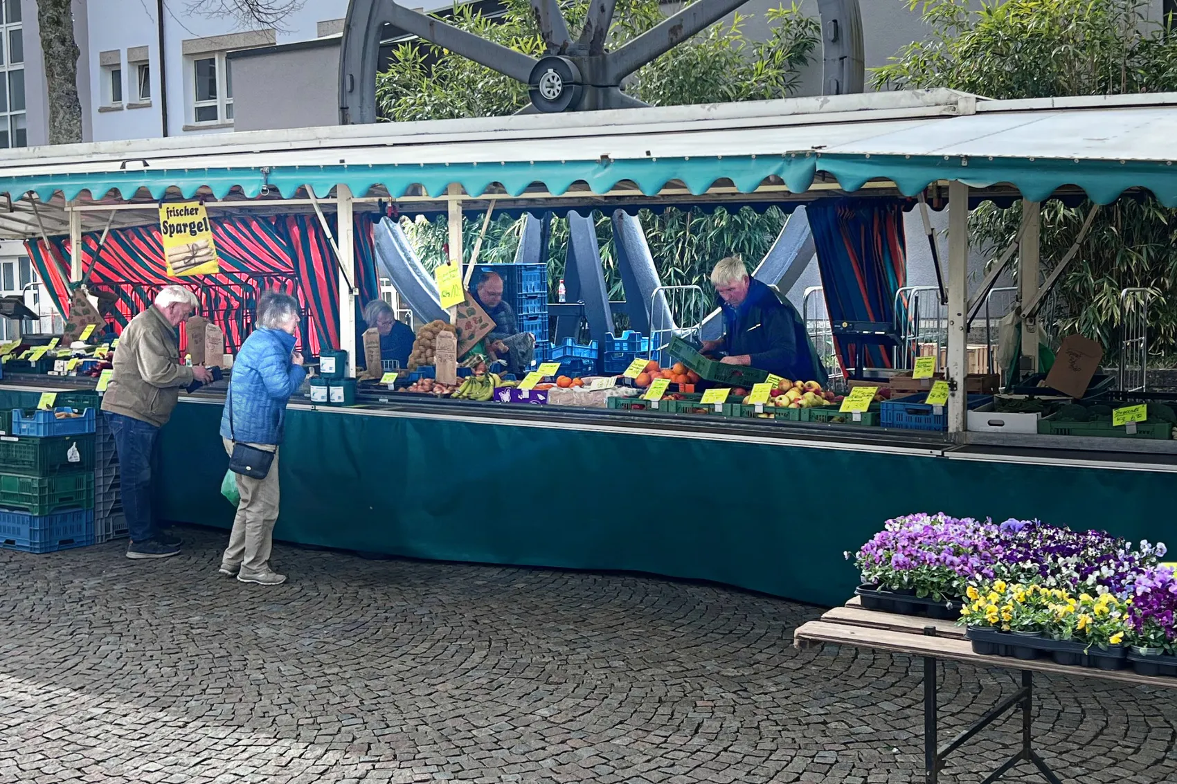 Gemüse- und Obsthändler Günter Deitelhoff bedient Kunden an seinem Marktstand in Plettenberg. Im Hintergrund steht seine Frau Heiderose. Vor dem Stand stehen Kisten mit Kartoffeln, Obst und Gemüse und ein Tisch mit blühenden Pflanzen.