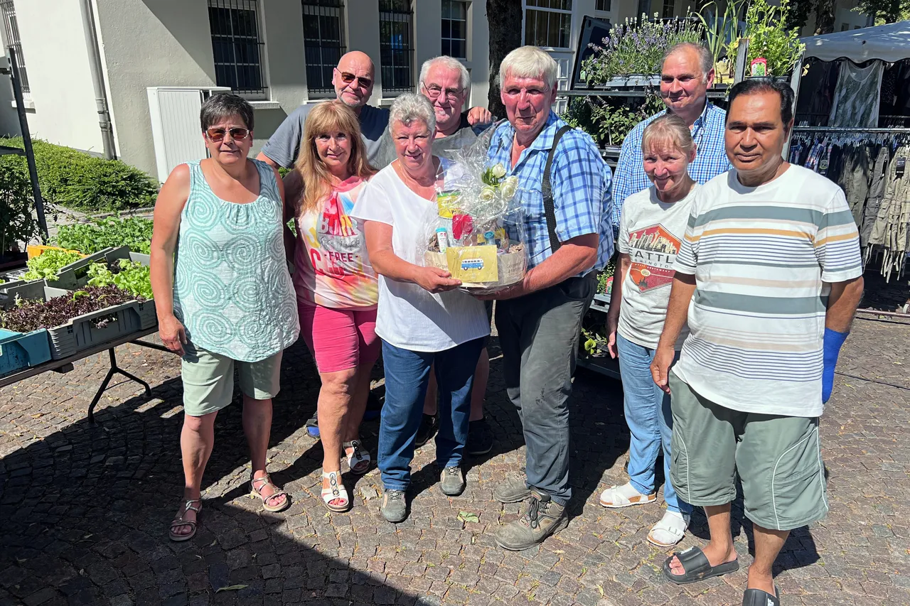 Eine Gruppe von sieben Personen steht lächelnd auf einem Pflasterweg vor Marktständen. In der Mitte halten zwei Männer ein Geschenk in den Händen.