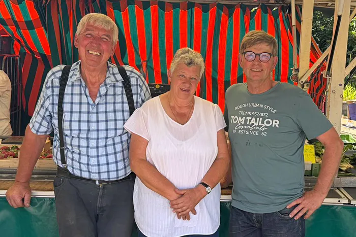 Drei Personen stehen lächelnd vor einem Marktstand mit Gemüse. Rechts trägt ein Mann ein hellgrünes T-Shirt mit Aufdruck, links ein blau-kariertes Hemd. Im Zentrum steht eine Frau mit weißem Oberteil.