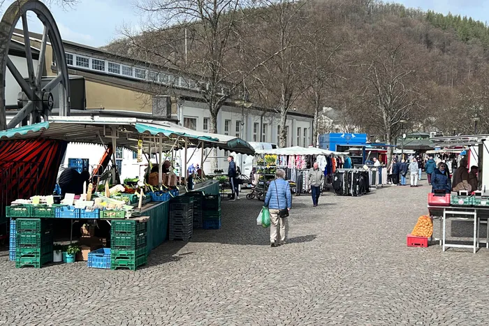 Ein belebter Wochenmarkt mit mehreren Ständen, auf denen Obst, Gemüse und Blumen angeboten werden. Im Vordergrund ein Stand mit Kartoffeln und Gemüse, während sich Besucher zwischen den Ständen bewegen.