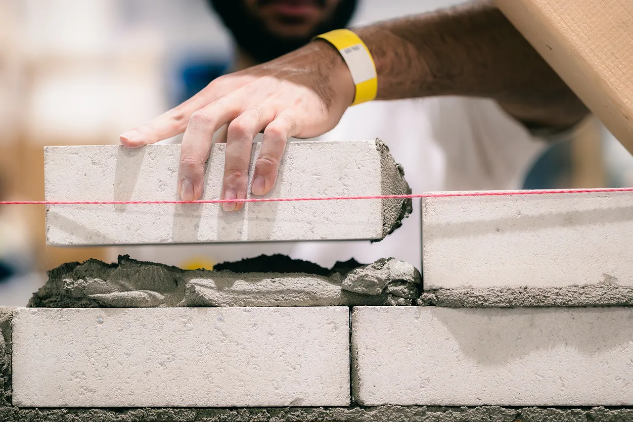 Eine Hand platziert einen hellen Ziegel auf eine mit Mörtel gefüllte Mauer, eine rote Schnur dient zur Ausrichtung.