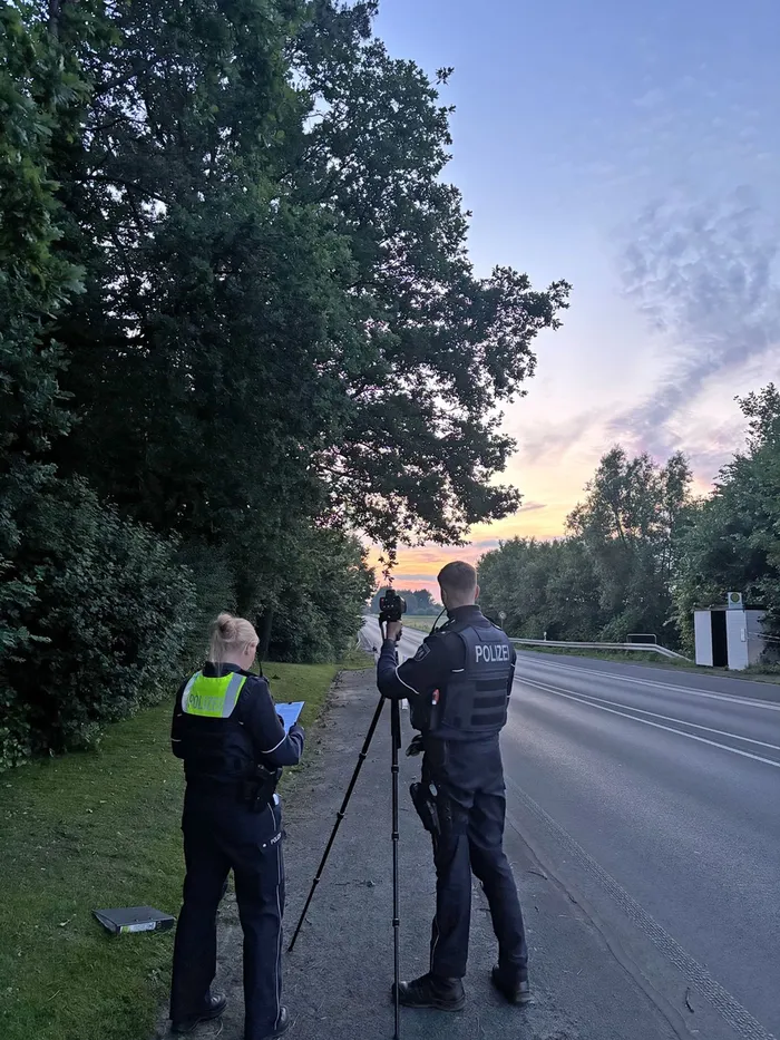 Zwei Polizisten stehen an einer Landstraße, einer bedient eine Messanlage, der andere filmt mit einer Kamera auf einem Stativ. Im Hintergrund ein Sonnenuntergang über Bäumen.