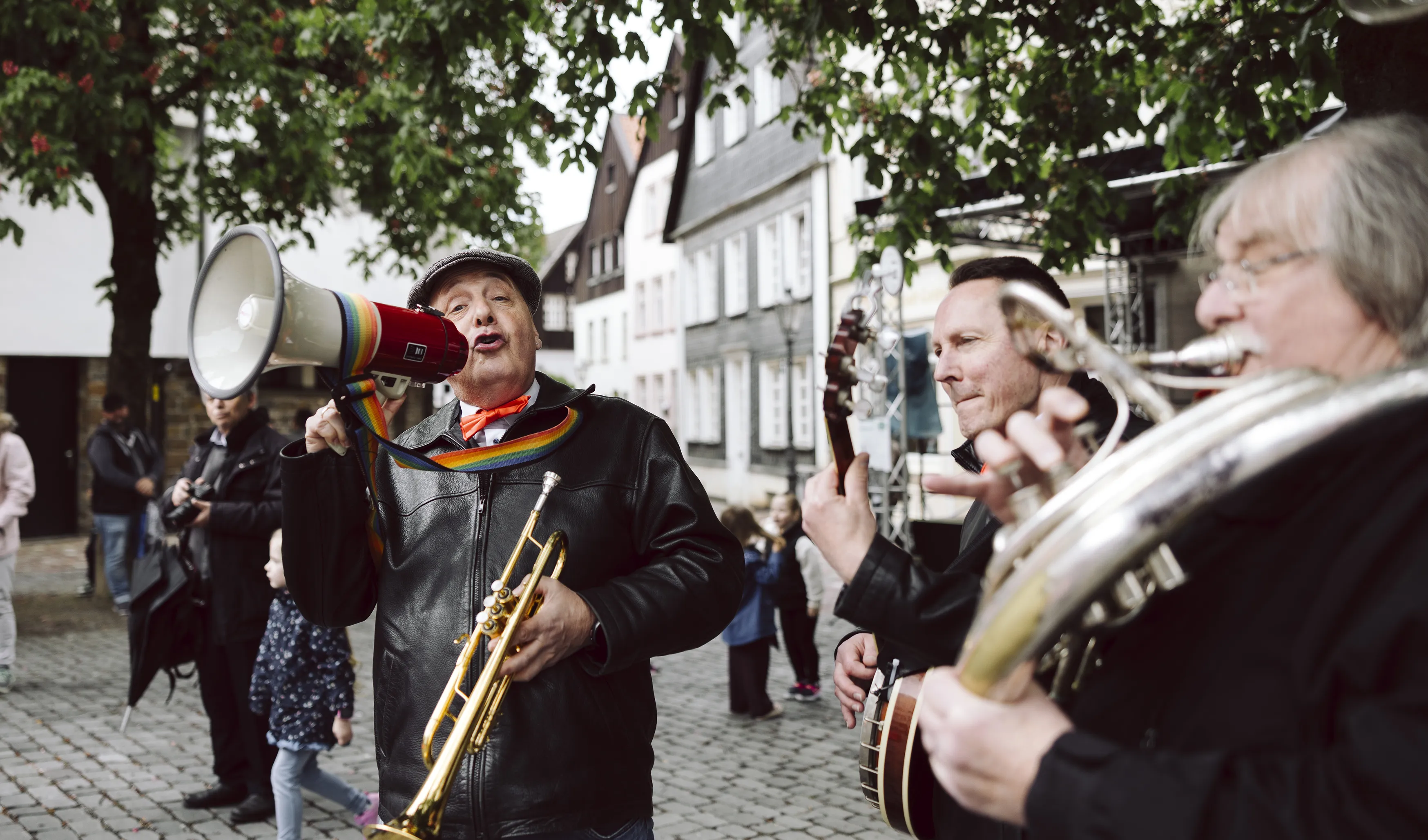 Eine Marching-Band spielt auf einer gepflasterten Straße in einer Altstadt. Ein Musiker mit Mütze und Trompete spricht in ein Megafon, während andere Bandmitglieder Posaune und Banjo spielen.