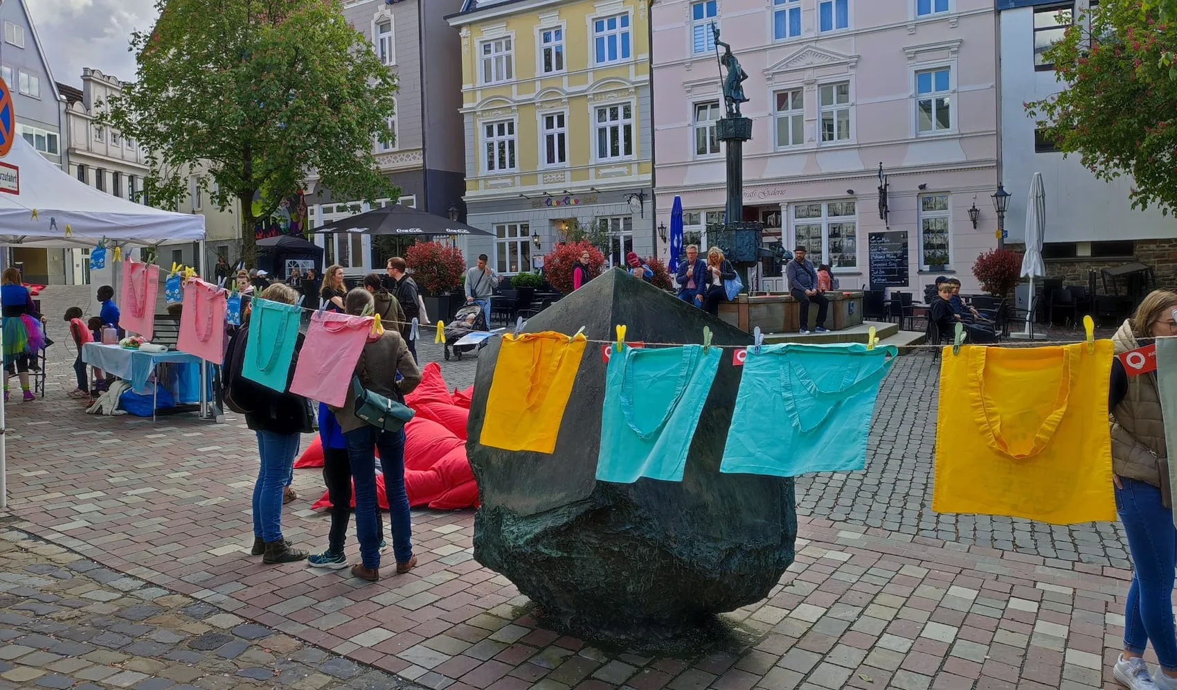 Bunte Tücher hängen an einer Skulptur auf dem Graf-Engelbert-Platz in Lüdenscheid, Menschen stehen und sitzen in der Nähe. Im Hintergrund sind Gebäude und Marktstände zu sehen.