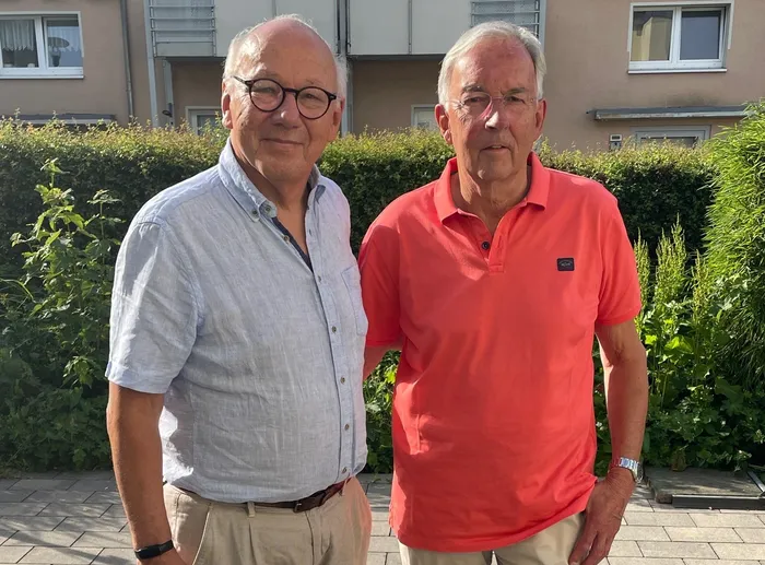 Zwei ältere Männer stehen nebeneinander vor Büschen und einem Wohnhaus. Links trägt der Mann ein helles Hemd und eine Brille, rechts ein rotes Poloshirt.