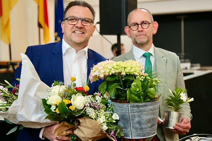 Zwei Männer im Anzug halten Blumen und eine blühende Pflanze in einem Eimer. Links der Bürgermeister, rechts der Gewählte.