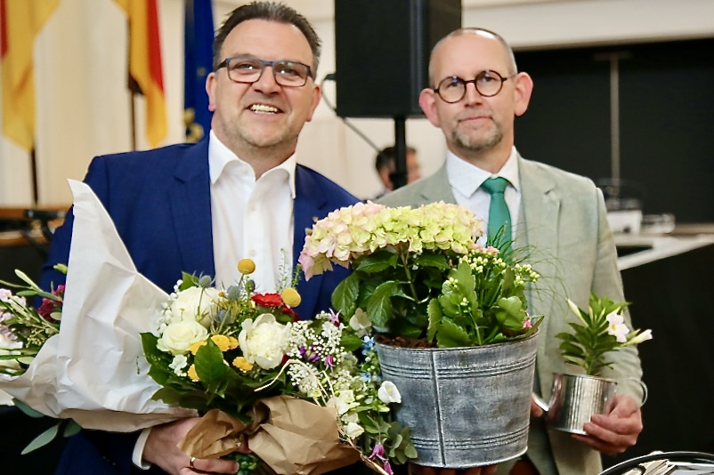 Zwei Männer im Anzug halten Blumen und eine blühende Pflanze in einem Eimer. Links der Bürgermeister, rechts der Gewählte.