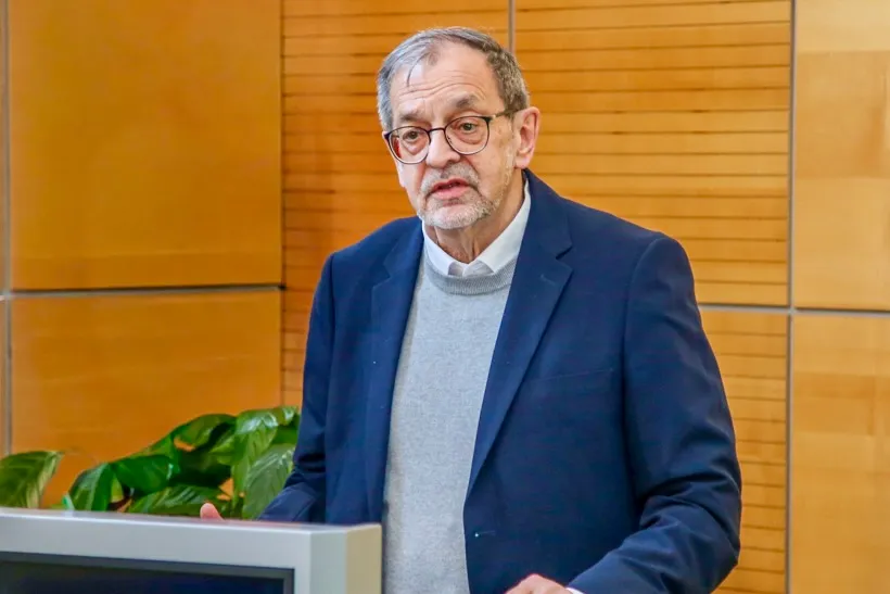 Michael Scheffler, ein älterer Mann mit Brille, spricht vor einem Pult. Er trägt einen blauen Blazer und einen grauen Pullover.