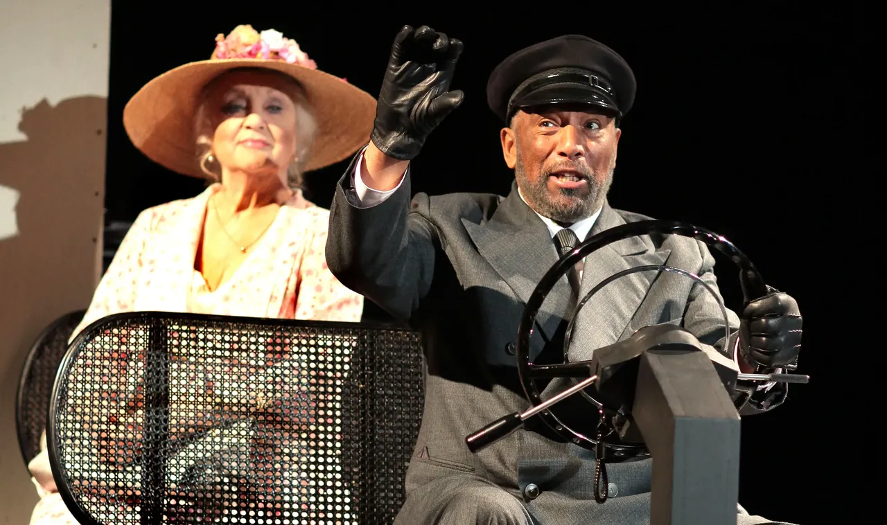 Doris Kunstmann und Ron Williams spielen in einer Bühneninszenierung von „Miss Daisy und ihr Chauffeur“ die Titelrollen. Williams hält als Chauffeur das Lenkrad eines Autos, während Kunstmann im Hintergrund sitzt und ihn beobachtet.