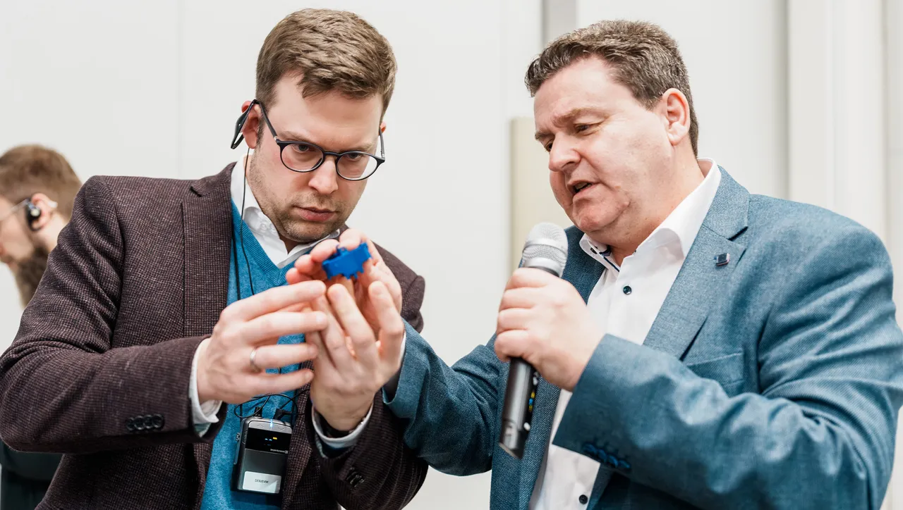 Zwei Männer betrachten gemeinsam ein kleines Gerät in der Hand, während einer von ihnen ein Mikrofon hält. Im Hintergrund sind weitere Personen zu sehen.