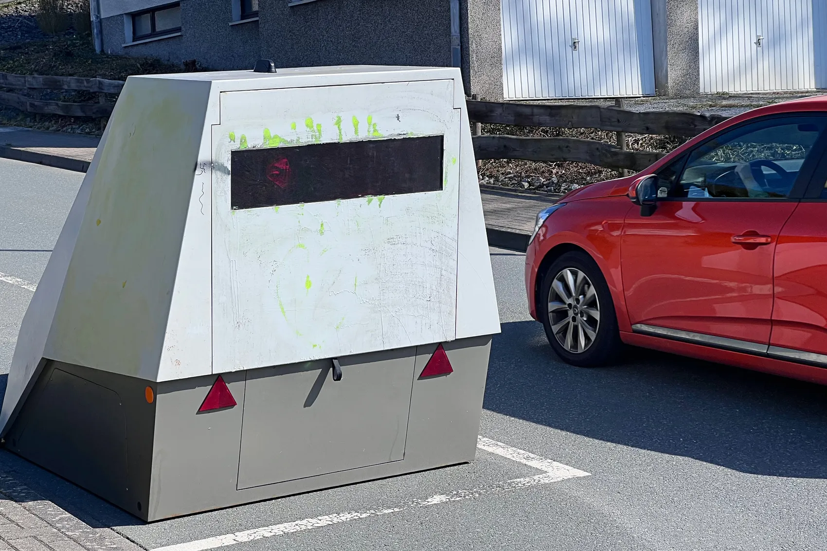 Grauer mobiler Blitzer mit roten Rückleuchten steht am Straßenrand neben einem roten Auto.