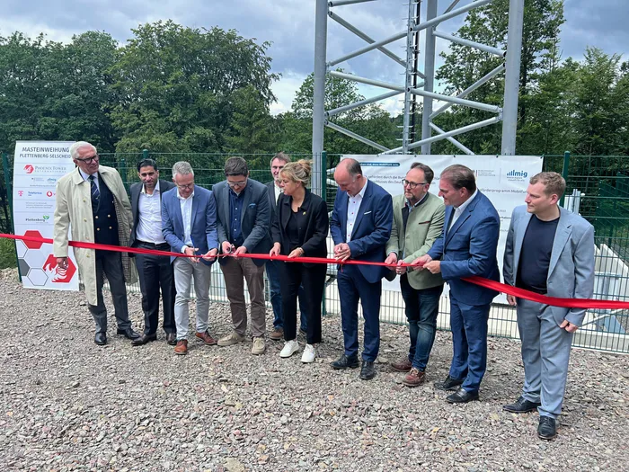 Eine Gruppe von Personen, darunter Mona Neubaur, schneidet ein rotes Band vor einem neuen Mobilfunkmast der Phoenix Tower Germany in Plettenberg-Selscheid durch. Im Hintergrund sind Banner mit den Logos der beteiligten Mobilfunkanbieter und der MIG zu sehen.