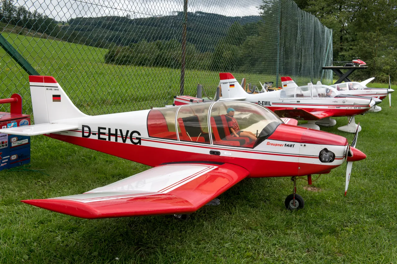Mehrere rot-weiße Modellflugzeuge stehen auf einer Rasenfläche vor einem grünen Zaun. Im Vordergrund ist ein Flugzeug mit der Kennung D-EHVG zu sehen.
