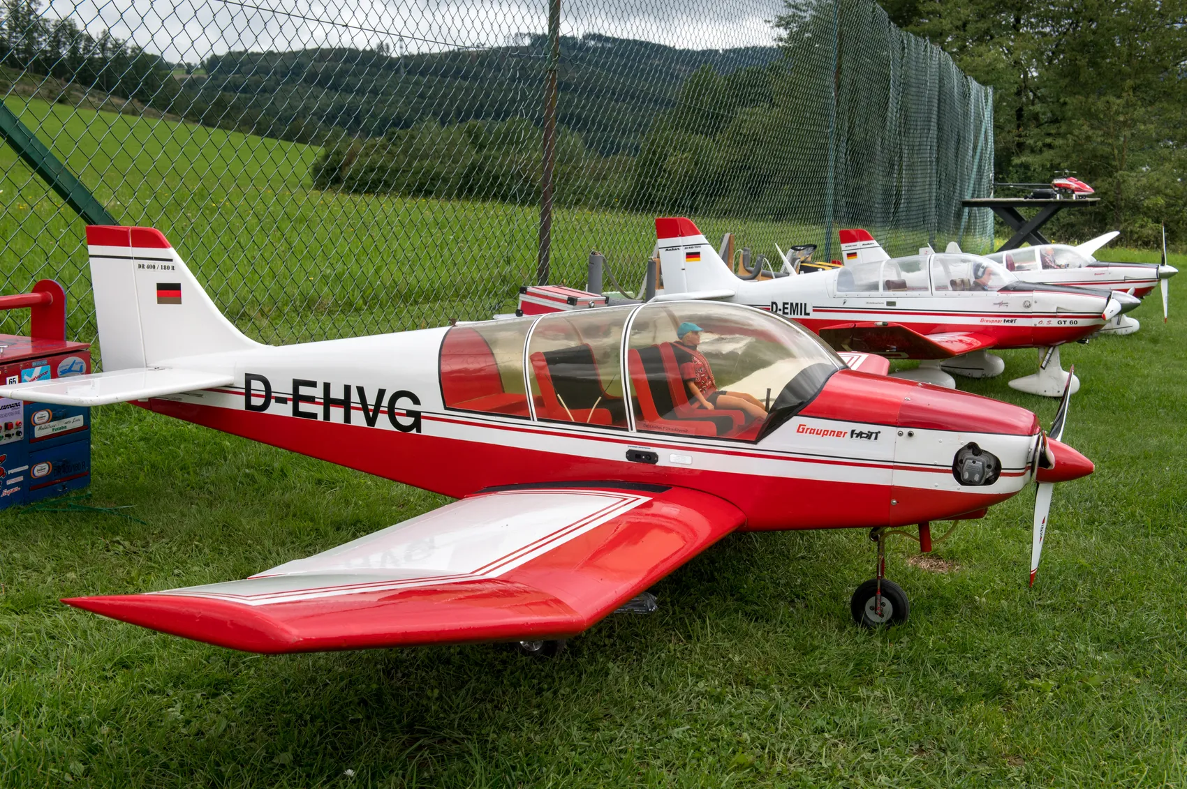 Mehrere rot-weiße Modellflugzeuge stehen auf einer Rasenfläche vor einem grünen Zaun. Im Vordergrund ist ein Flugzeug mit der Kennung D-EHVG zu sehen.
