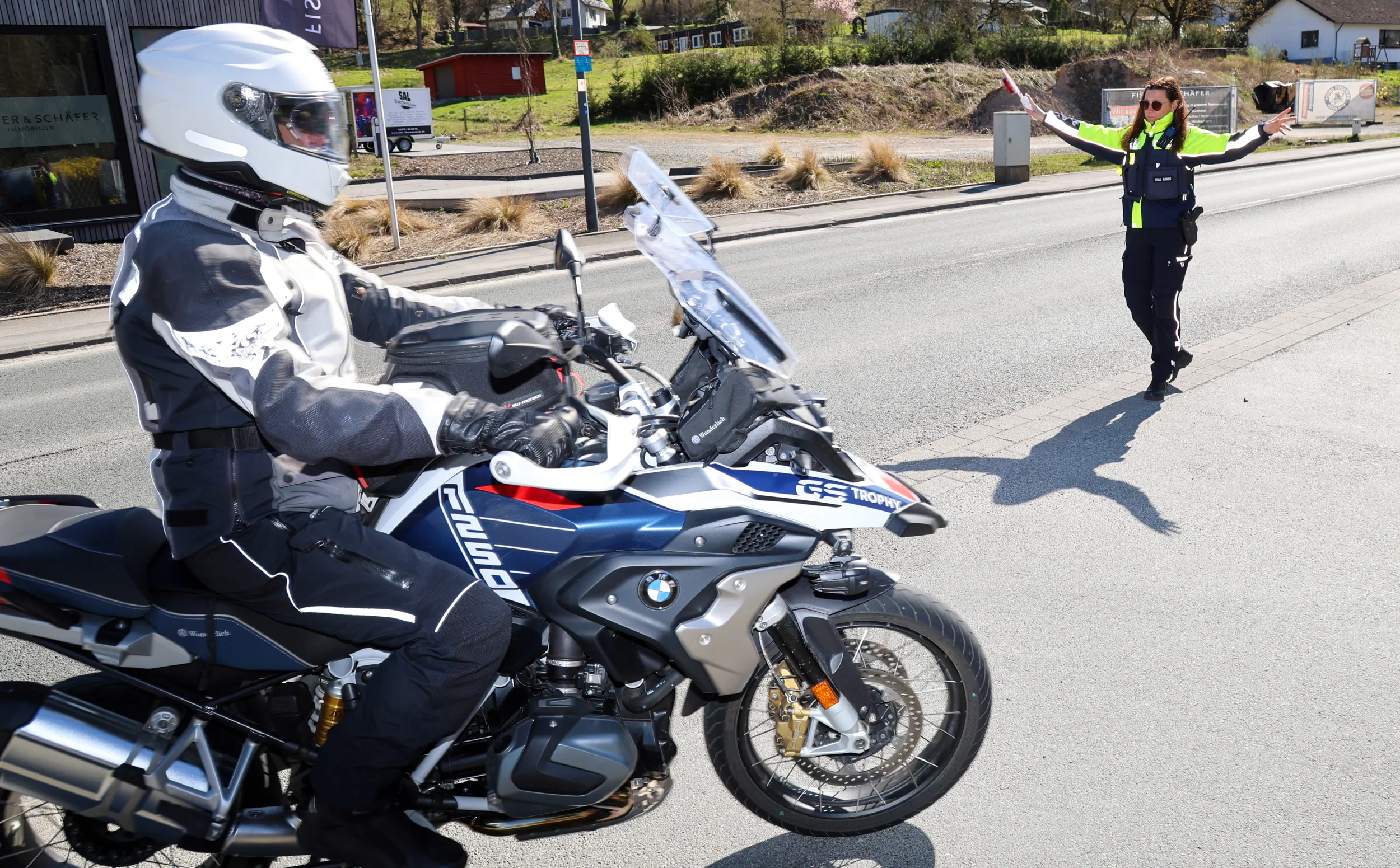 Motorradfahrer in Schutzkleidung wird von einer Polizistin auf einer Landstraße kontrolliert. Im Hintergrund ist ein Gebäude und ein DRK-Fahrzeug zu sehen.
