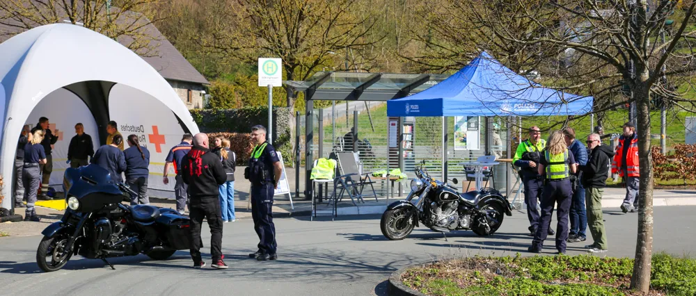 Polizisten kontrollieren Motorradfahrer auf einem Parkplatz, während ein rotes Zelt des Deutschen Roten Kreuzes im Hintergrund steht.
