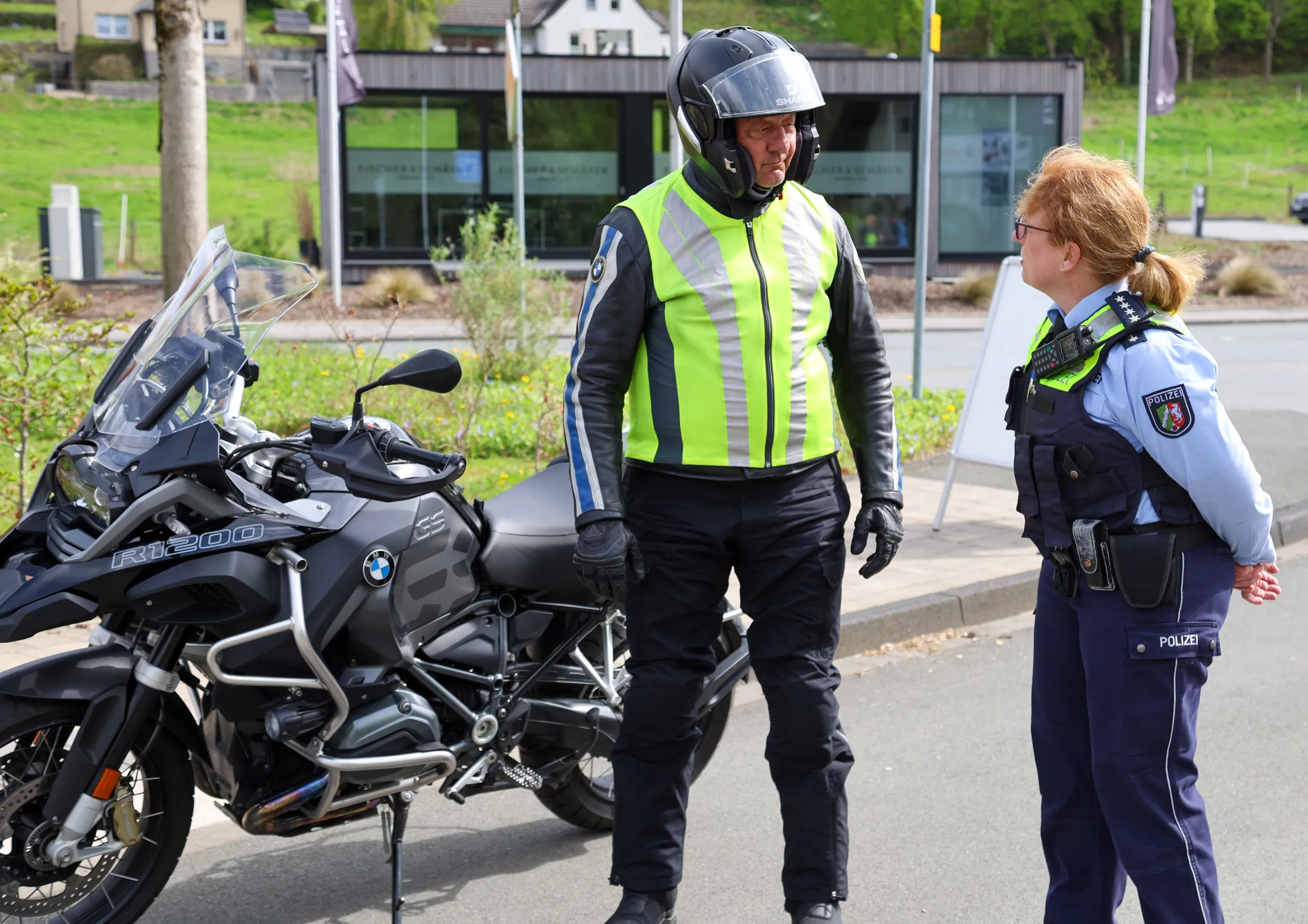 Polizistin spricht Motorradfahrer in voller Ausrüstung an. Im Hintergrund ein Gebäude und ein gepflegter Grünstreifen.