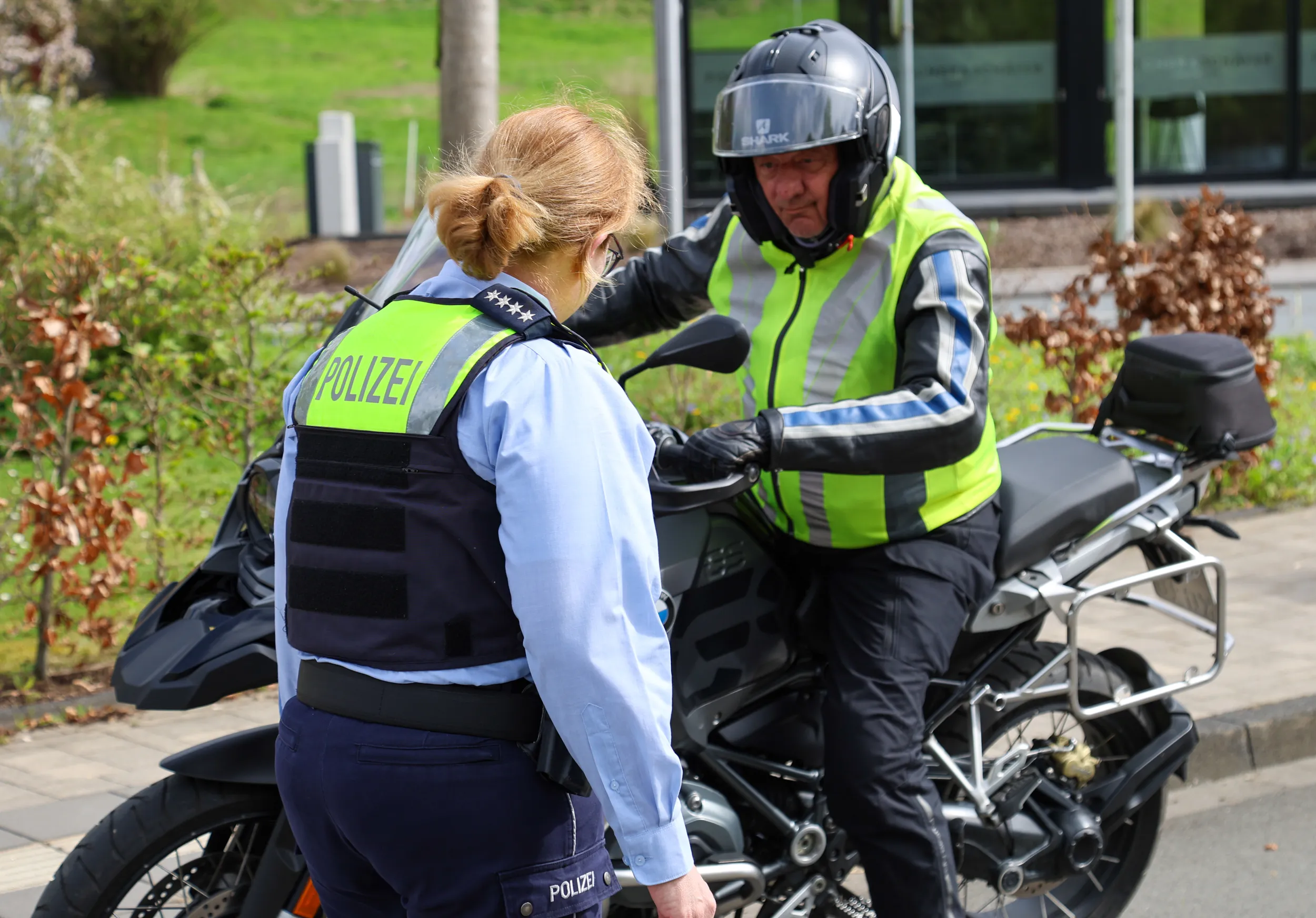 Polizistin im Gespräch mit einem Motorradfahrer während einer Verkehrskontrolle. Im Hintergrund ein Gebäude und herbstliche Vegetation.