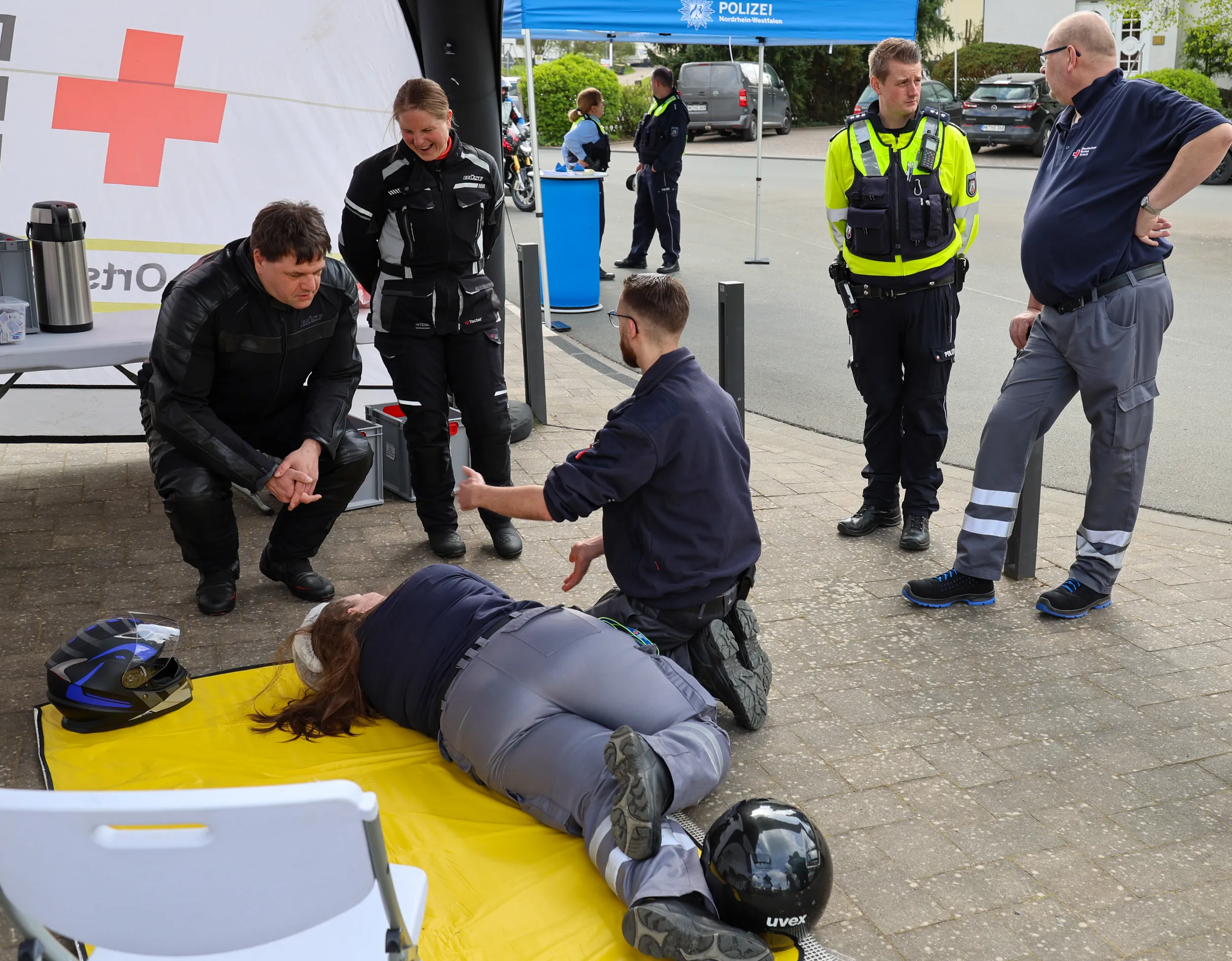 Zwei Sanitäter des Deutschen Roten Kreuzes versorgen eine Person, die auf einer gelben Rettungsdecke liegt. Im Hintergrund stehen Polizisten und ein Zelt mit dem DRK-Logo.