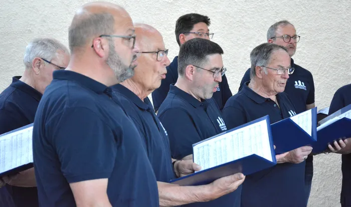 Eine Gruppe von Männern singt im Freien, alle halten Notenblätter in der Hand. Sie tragen dunkle Poloshirts mit einem Logo auf der Brust.