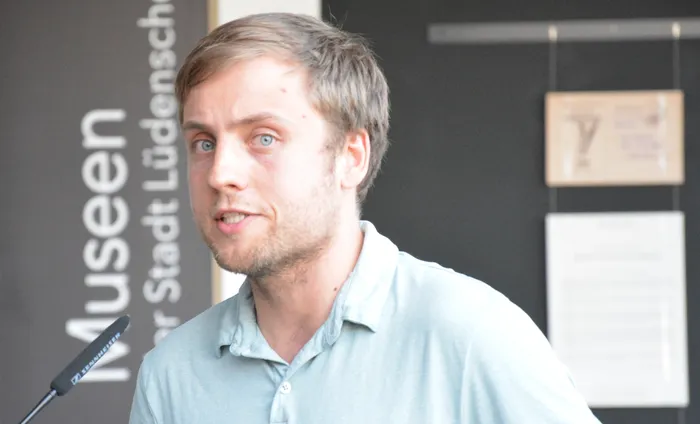Ein Mann mit blonden Haaren und Bart spricht in ein Mikrofon. Im Hintergrund ist ein Banner mit der Aufschrift 