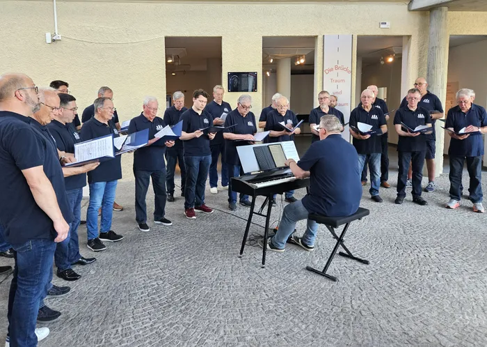 Ein Chor aus etwa 20 Männern steht vor einem hellen Gebäude und singt Notenblätter. Ein Mann spielt auf einem Keyboard. Im Hintergrund ist ein Banner mit der Aufschrift „Die Brücke. Traum und Alptraum“ zu sehen.