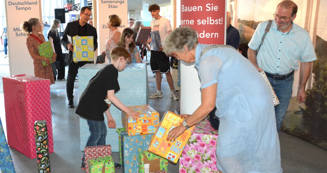 Kinder und eine ältere Frau stellen bunte, geschenkt verpackte Pakete als Teil einer Brückenkonstruktion auf. Im Hintergrund weitere Besucher der Ausstellungseröffnung.