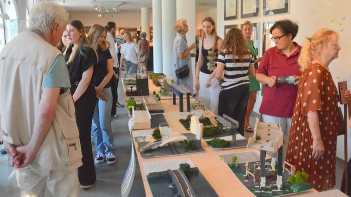 Menschen betrachten Architekturmodelle in einer hellen Ausstellungshalle. Die Modelle zeigen Stadtansichten und Brückenkonstruktionen.
