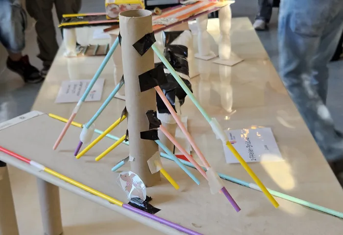 Ein selbstgebautes Modell einer Brücke aus Pappe und Stäben steht auf einem Tisch. Im Hintergrund sind weitere Ausstellungsstücke und Besucher zu erkennen.