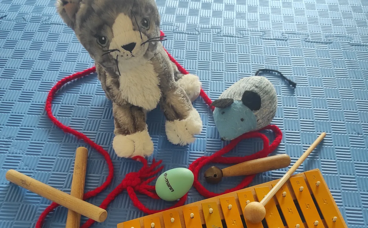 Ein Plüschkater mit roter Schleife sitzt neben einer Stoffmaus, einem bemalten Osterei und einem Xylophon mit zwei Schlägeln auf einer blauen Sportmatte.