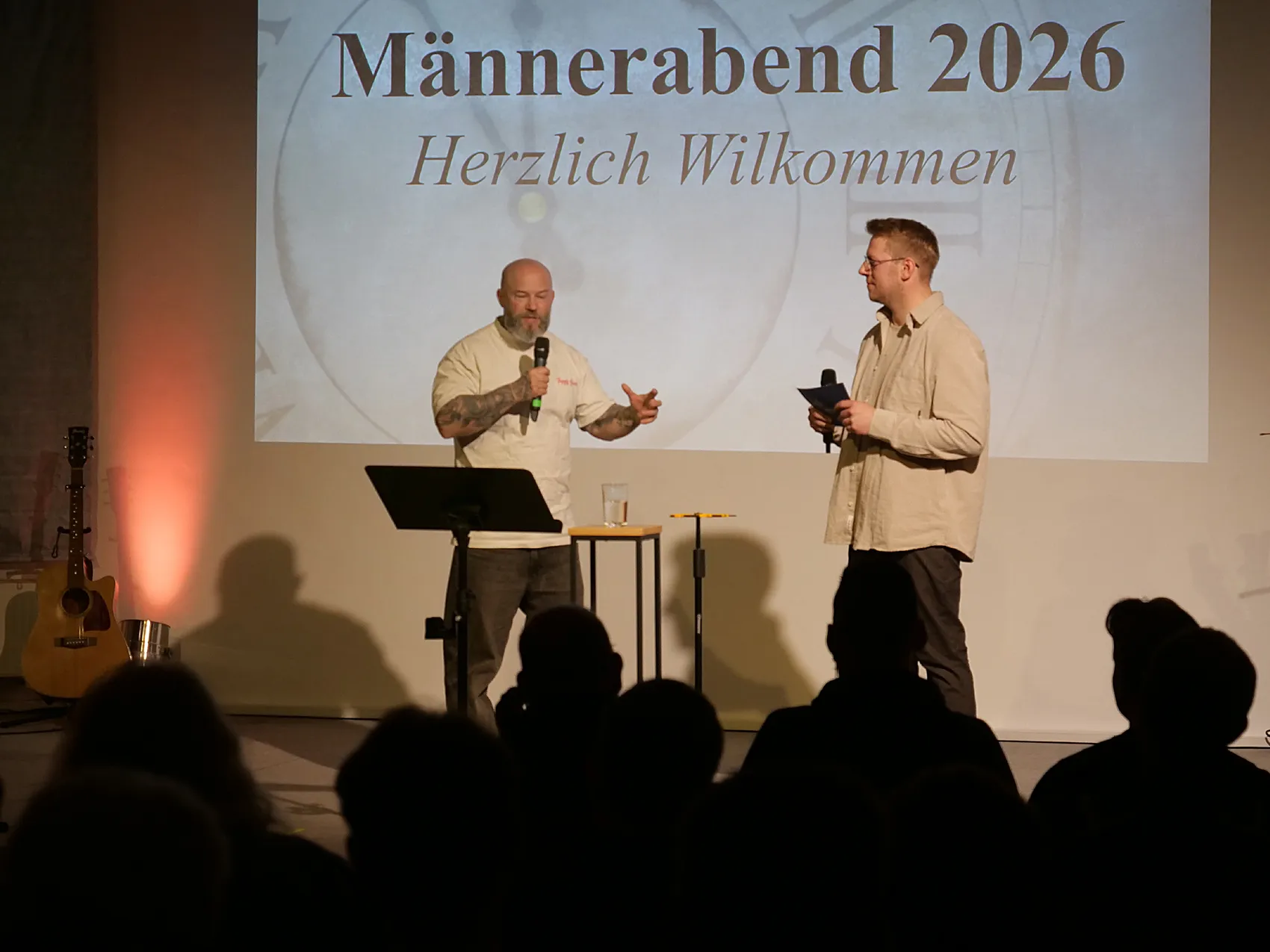 Zwei Männer auf einer Bühne sprechen vor einem Publikum. Im Hintergrund steht die Aufschrift „Männerabend 2026 Herzlich Willkommen“.