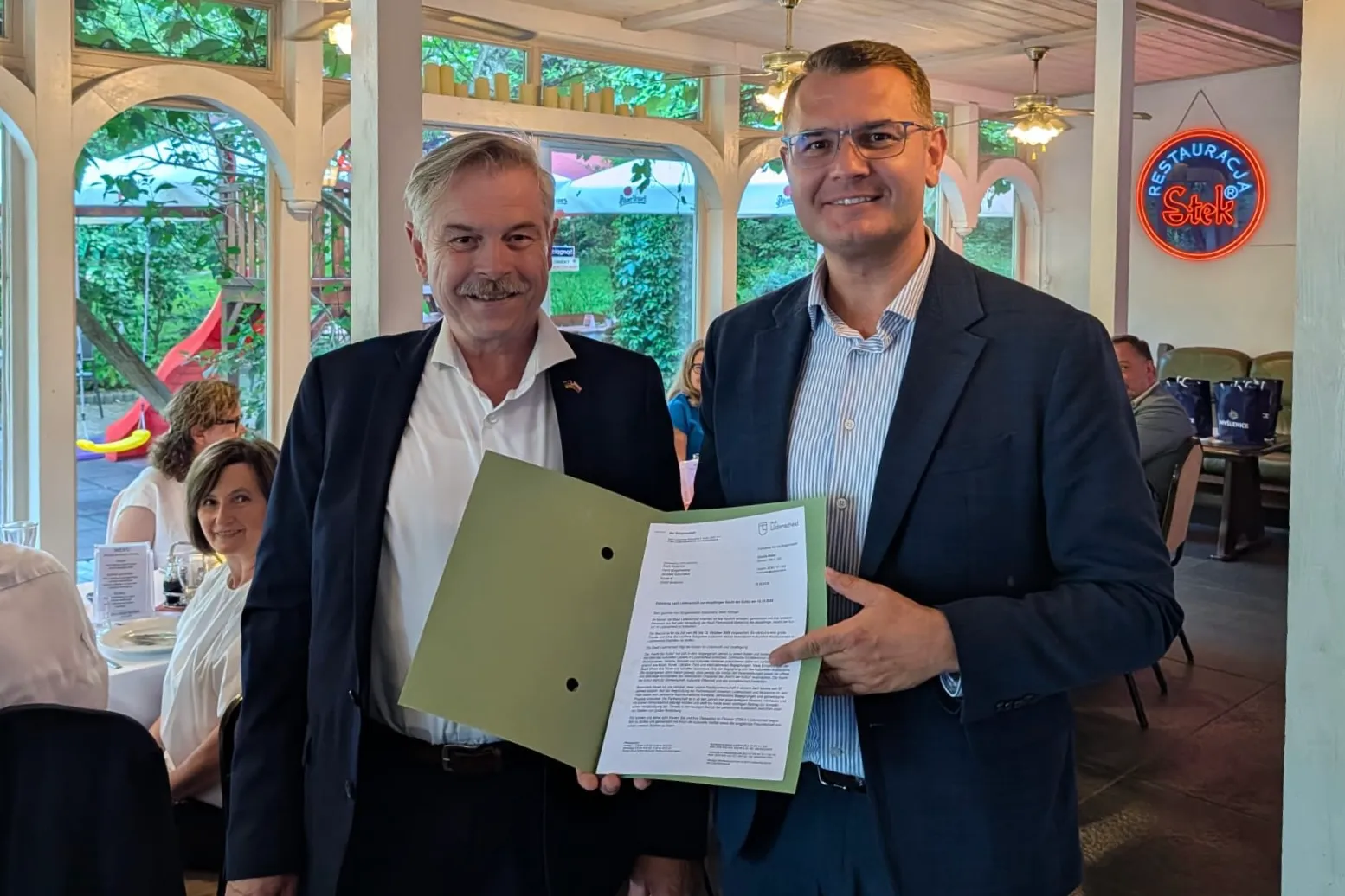 Zwei Männer in Anzügen stehen sich lächelnd gegenüber und halten ein Dokument. Im Hintergrund sind weitere Personen und ein Restaurant zu sehen.
