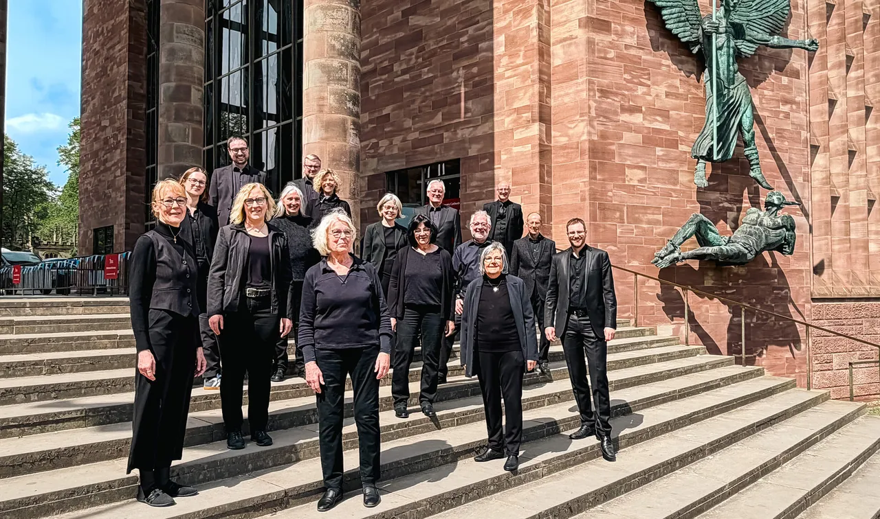 Gruppe von etwa 15 Personen in dunkler Kleidung posiert auf den Stufen vor einer Kathedrale mit einer großen Engelsstatue.