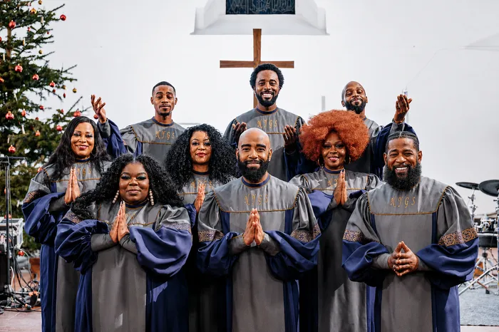 Die Mitglieder des Chores New York Gospel Stars posieren in grauen und blauen Roben vor einem Weihnachtsbaum und einem Holzkreuz.