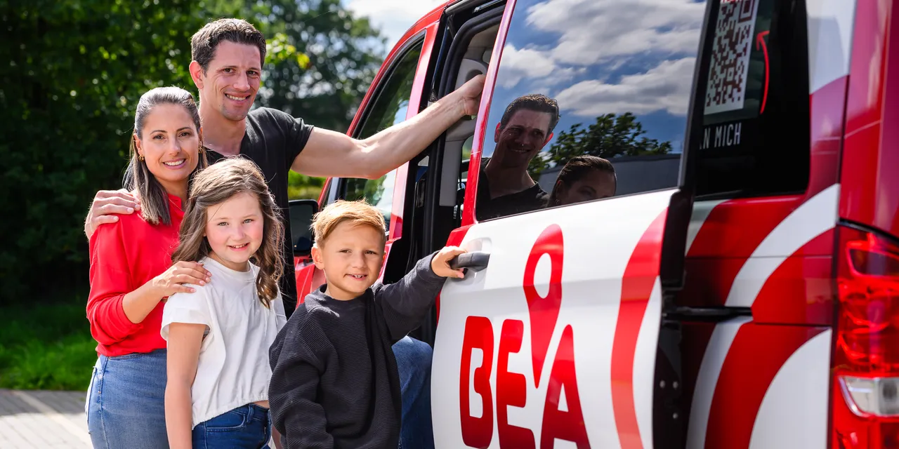Familie steht vor einem roten Kleinbus mit dem Aufdruck „BEA“ und einem QR-Code. Ein Mann zeigt auf den Bus, während seine Frau und zwei Kinder lächeln. Im Hintergrund sind weitere Personen im Bus zu sehen.