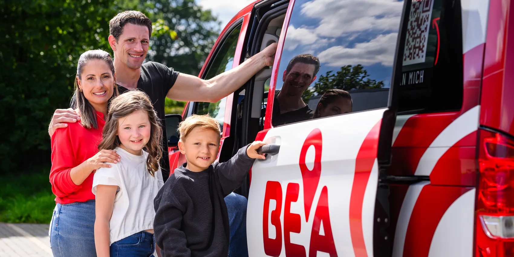 Familie steht vor einem roten Kleinbus mit dem Aufdruck „BEA“ und einem QR-Code. Ein Mann zeigt auf den Bus, während seine Frau und zwei Kinder lächeln. Im Hintergrund sind weitere Personen im Bus zu sehen.