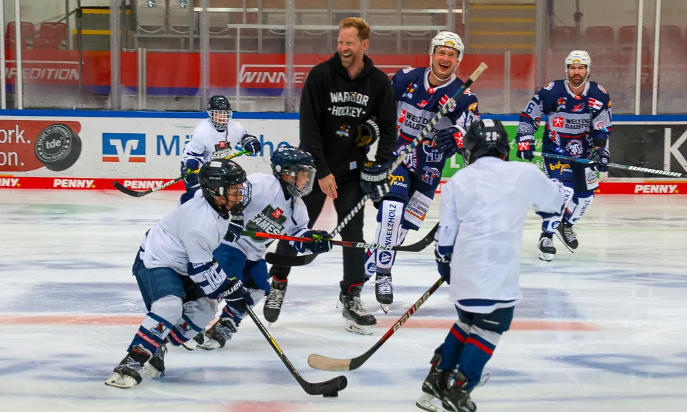 Eishockeytrainer Lasse Kopitz lacht mit jungen Spielern auf dem Eis, während diese mit Schlägern und Puck posieren.