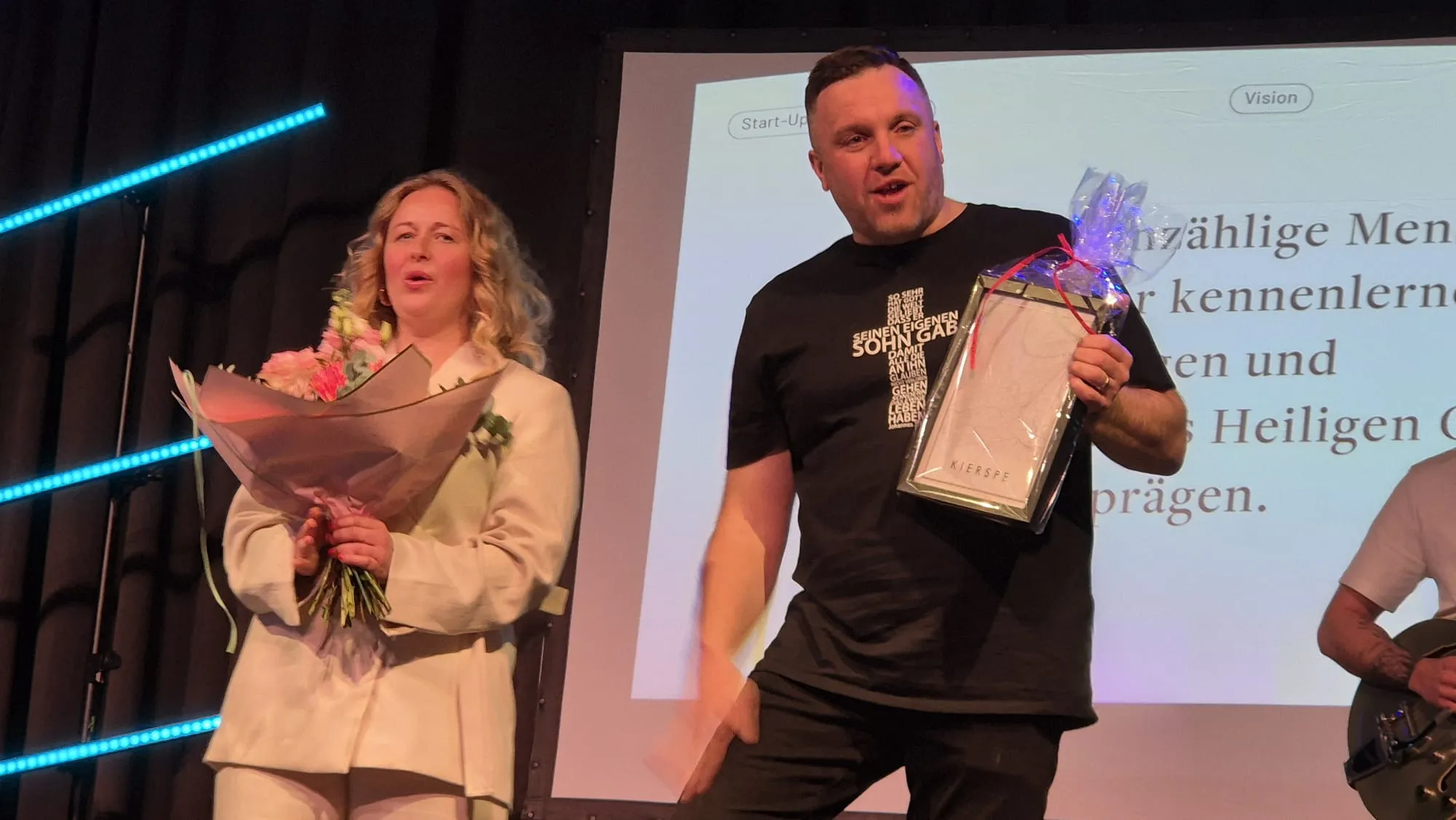 Mann und Frau stehen auf einer Bühne, der Mann hält eine Auszeichnung und einen Blumenstrauß. Im Hintergrund ist ein Bildschirm mit Text zu sehen.