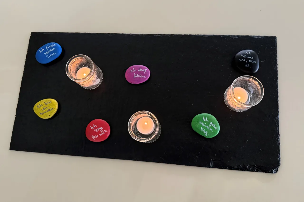Bunte Steine mit handgeschriebenen Texten und brennende Teelichter auf einer schwarzen Schieferplatte.