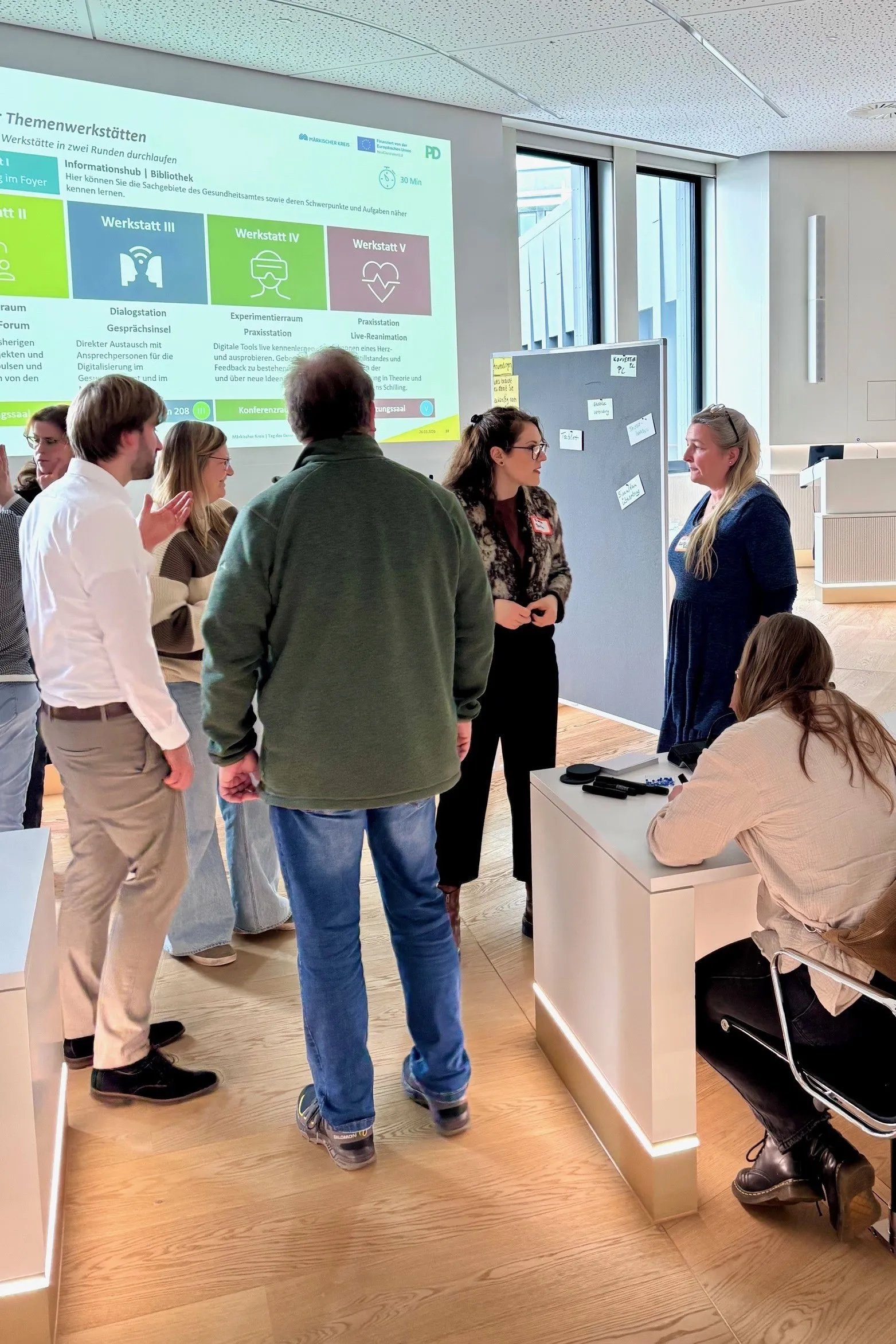 Gesprächsgruppe vor Plakaten mit den Überschriften „Werkstatt III“ bis „Werkstatt V“ auf einem Netzwerktreffen zum Thema Digitalisierung im Gesundheitsdienst.