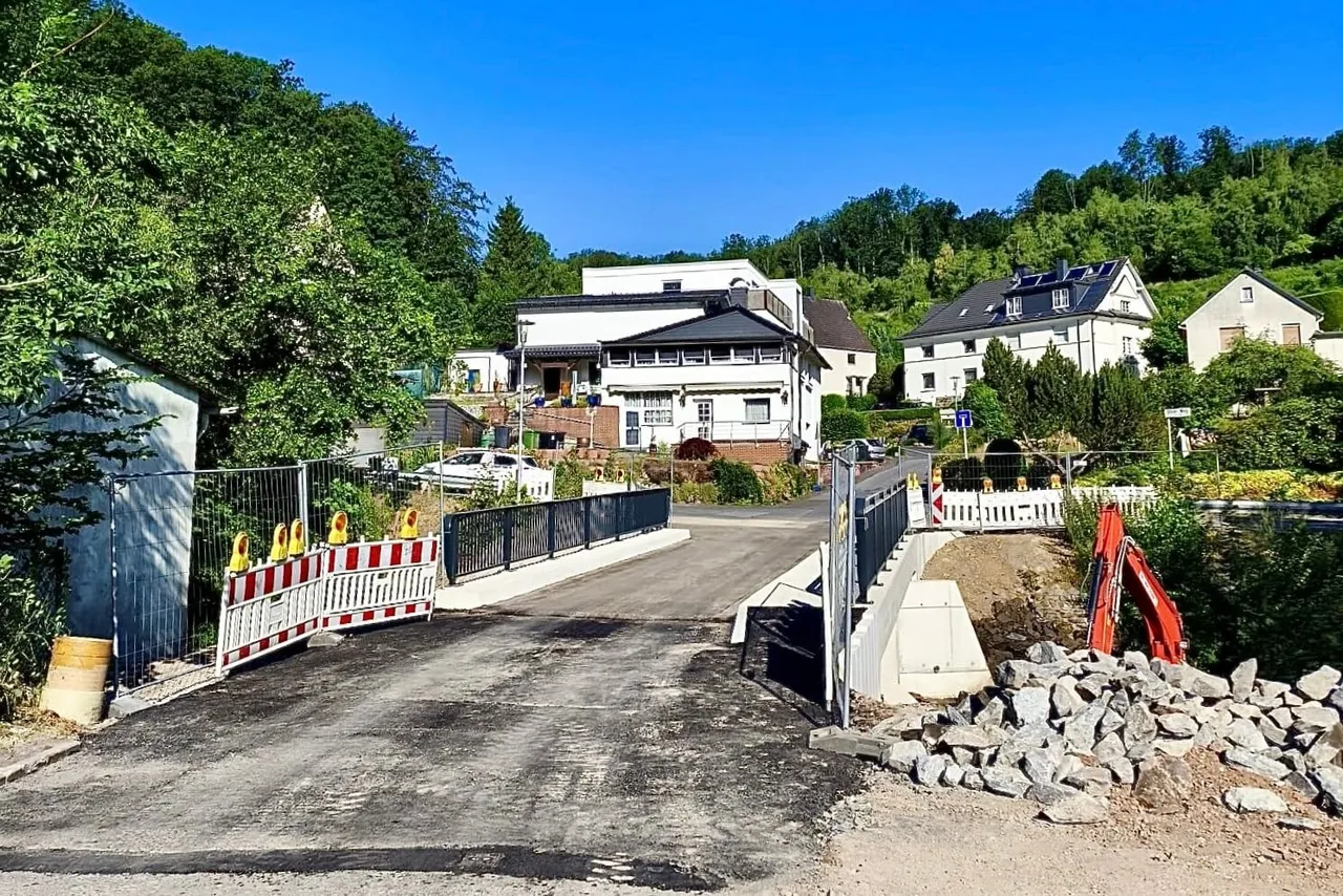 Die neue Brücke über die Volme ist für den Verkehr freigegeben, nachdem die alte Brücke an der „Schiefen Ahelle“ ersetzt wurde. Im Vordergrund sind Absperrungen und Bauarbeiten zu sehen, im Hintergrund Wohnhäuser und Bäume.